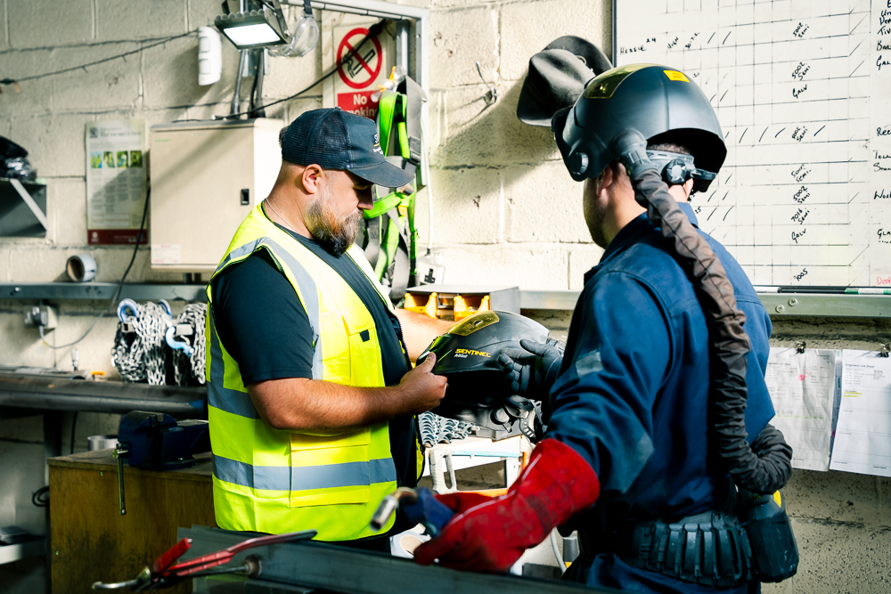 Technician in high-visibility vest talking to a welder in a workshop.