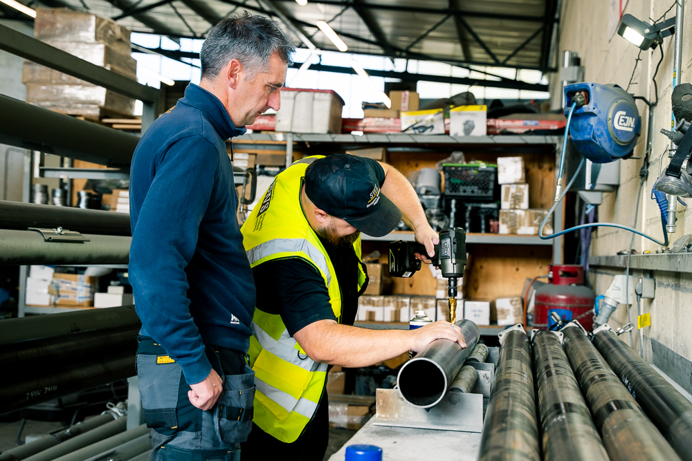 Technician drilling metal pipe while colleague watches in workshop.