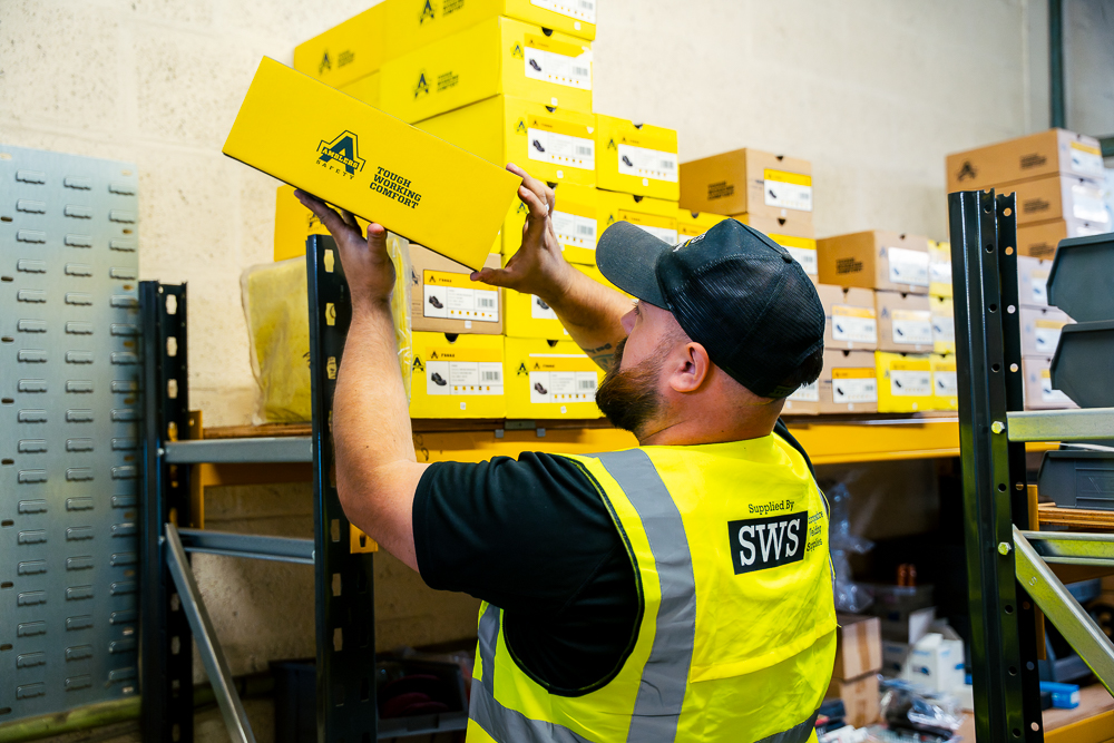 SWS worker in high-visibility vest lifting yellow boxes in a warehouse.