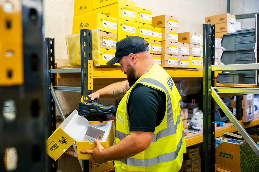 Warehouse worker lifting a yellow PPE shoes and box from storage shelves.
