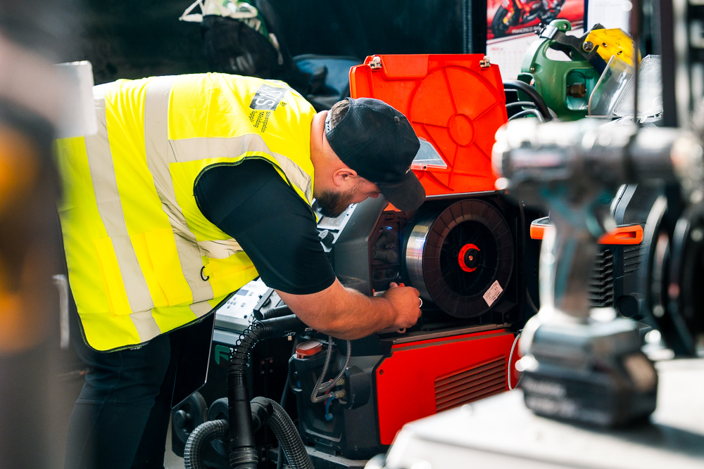 Technician drilling metal pipe while colleague watches in workshop.